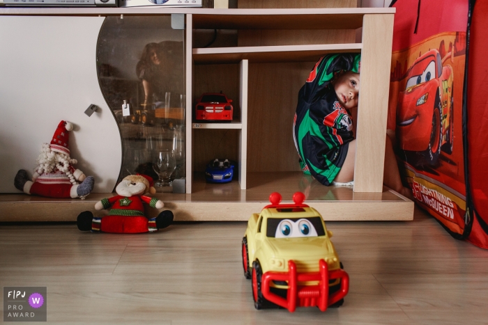 A little boy sits hiding on a shelf surrounded by toy cars in this photo by a Rio Grande do Sul, Brazil family photojournalist. 