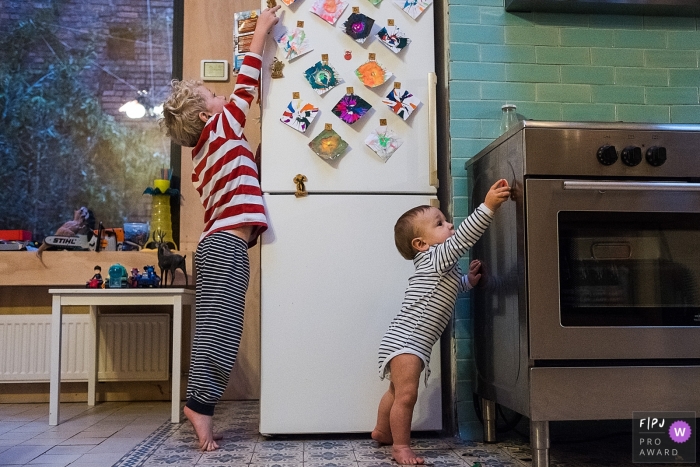 A young boy and his baby brother play with magnets on the refrigerator and stove in this photo by an Antwerpen family photojournalist. 