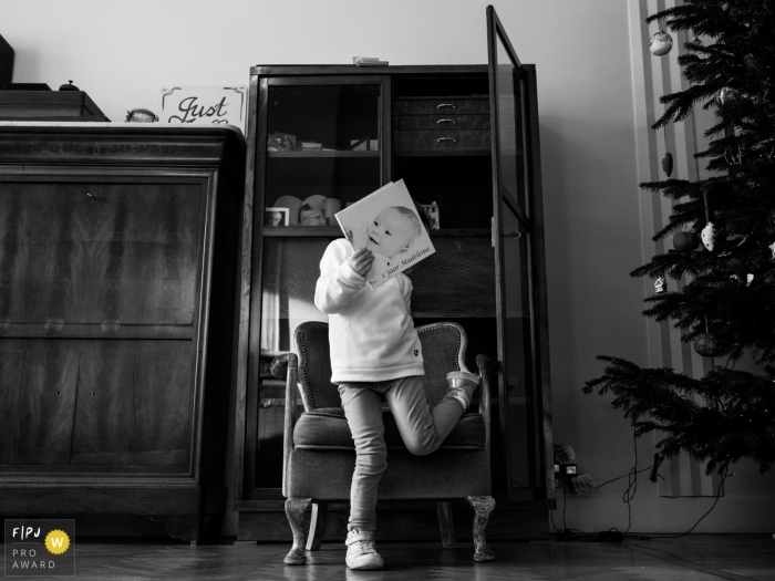 Photo by: <a href="https://www.fpja.com/documentary-family-photography-contests/joshua-dhondt/antwerpen/2075196">Joshua Dhondt - Belgium</a> A child covers their face with a book with a large baby face on the cover in this black and white photo by an Antwerpen family photojournalist.