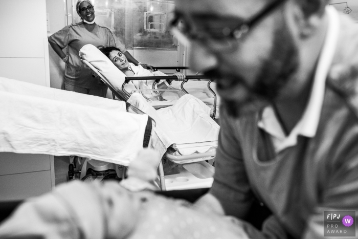 A mother looks across the hospital room where her husband hods their newborn in this black and white photo by a Sao Paulo birth photographer.