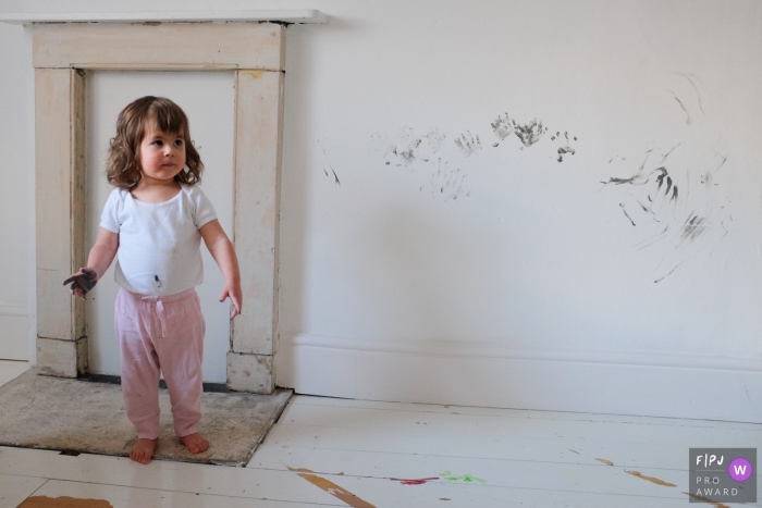 A little girl stands next to a white wall covered in hand prints with paint on her hands in this photo by a Gloucestershire, England family photojournalist.