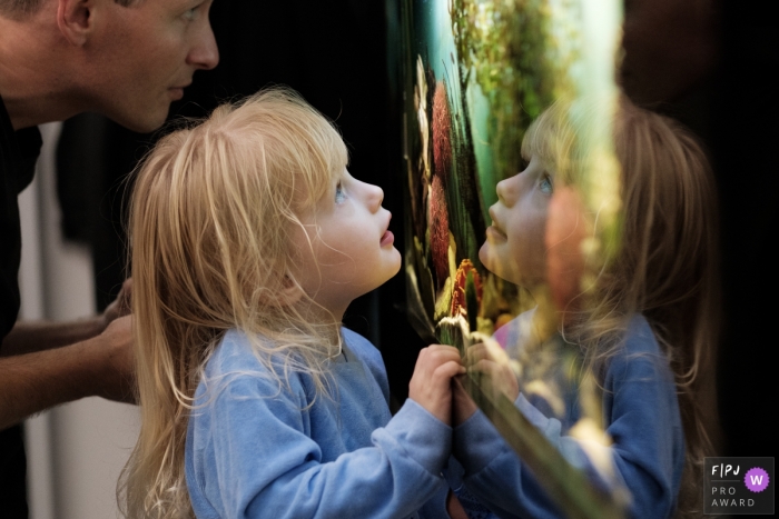 Photo by: <a href="https://www.fpja.com/documentary-family-photography-contests/jennifer-adams/gloucestershire/2075981">Jennifer Adams - United Kingdom</a> A little girl's face is reflected in a window as she looks outside with her father in this photo by a Gloucestershire, England family photojournalist.