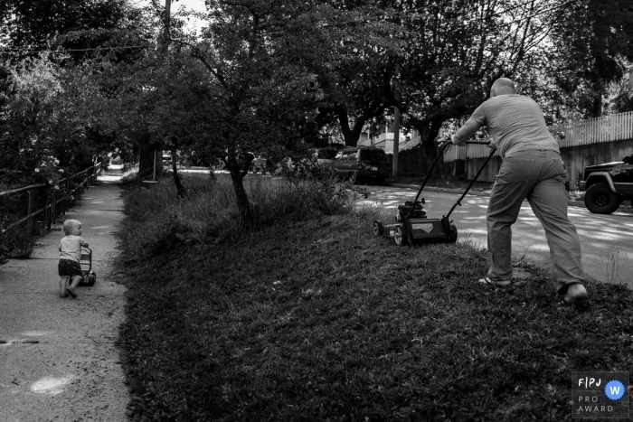 Photo by: <a href="https://www.fpja.com/documentary-family-photography-contests/bobbi-barbarich/british-columbia/2076566">Bobbi Barbarich - Canada</a> A little boy copies his father mowing the lawn as he pushes his own toy mower in this black and white photo by a British Columbia, Canada family photojournalist.