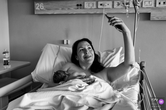 Photo by: <a href="https://www.fpja.com/documentary-family-photography-contests/lavinia-nitu/modena/2076676">Lavinia Nitu - Italy</a> A mom takes a selfie in the hospital as she holds her newborn baby in this black and white photo by a Modena birth photographer.
