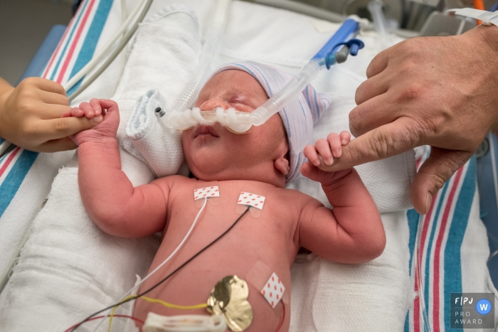 A newborn hooked up to monitors in the hospital holds its parents' fingers in each hand in this photo by a Connecticut birth photographer.