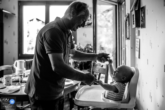 Un homme joue avec un bébé assis sur une chaise haute dans cette photographie en noir et blanc réalisée par un photographe de famille documentaire savoyard