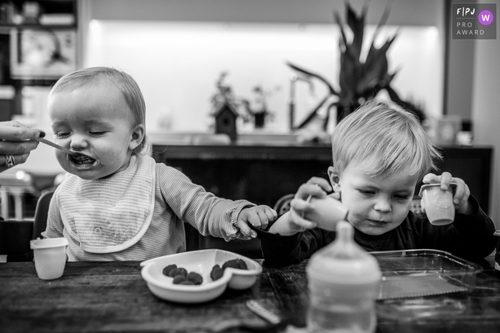 Photo by: <a href="https://www.fpja.com/documentary-family-photography-contests/nadine-court/savoie/2078056">Nadine Court - France</a> A little boy plays with yogurt cups as his mom feeds his baby brother in this black and white photo by a Savoie family photojournalist.