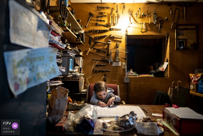 Photo by: <a href="https://www.fpja.com/documentary-family-photography-contests/romulo-morishita/madison/2078281">Romulo Morishita - United States</a> A boy writes on a table calendar in an office with tools covering the walls in this photo by a Madison, WI family photojournalist.