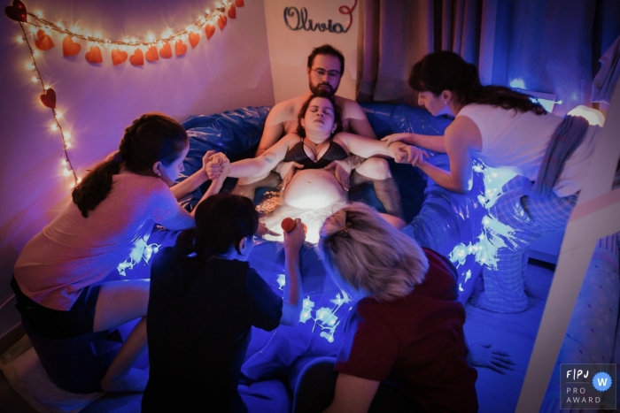 Two girls hold a woman's hands as she sits in a birthing tub with her husband in this photo by a Manaus birth photographer. 