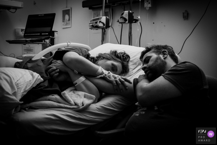 A wife holds her husbands arm as she lays in a hospital bed and the two of them sleep in this black and white photo by a Drenthe, Netherlands birth photographer.