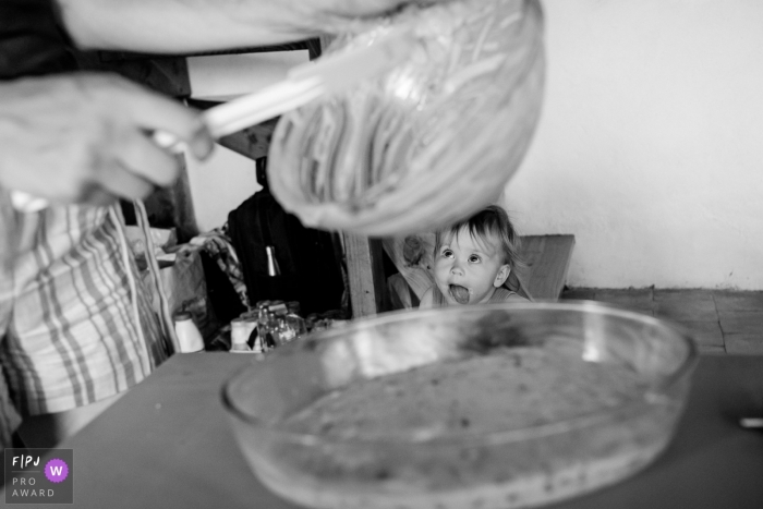 A little girl with her tongue sticking out watches her mother make food in the kitchen in this black and white photo by a Nantes family photojournalist. 