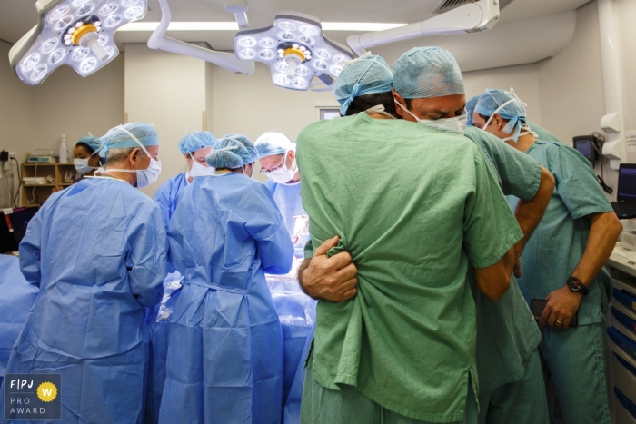Photo by: <a href="https://www.fpja.com/documentary-family-photography-contests/graziela-ventura/s%C3%A3o-paulo/2080011">Graziela Ventura - Brazil</a> São Paulo hospital birth photographer captured this photo of two men in scrubs hugging while a hospital team operates in the background
