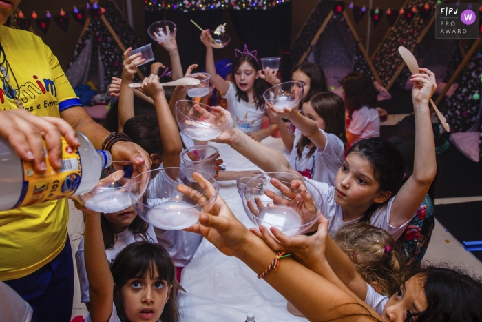 Photo by: <a href="https://www.fpja.com/documentary-family-photography-contests/graziela-ventura/s%C3%A3o-paulo/2080101">Graziela Ventura - Brazil</a> Several children hold up their cups to get refills as they sit around along table in this photographic image by a Sao Paulo, Brazil family photojournalist.