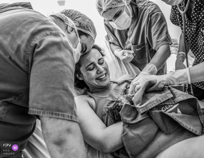 Photo by: <a href="https://www.fpja.com/documentary-family-photography-contests/alessandra-corveloni/rio-de-janeiro/2080606">Alessandra Corveloni - Brazil</a> A mother holds her newborn in the hospital surrounded by her husband and nurses in this black and white birthing photograph by a Rio de Janeiro photographer.