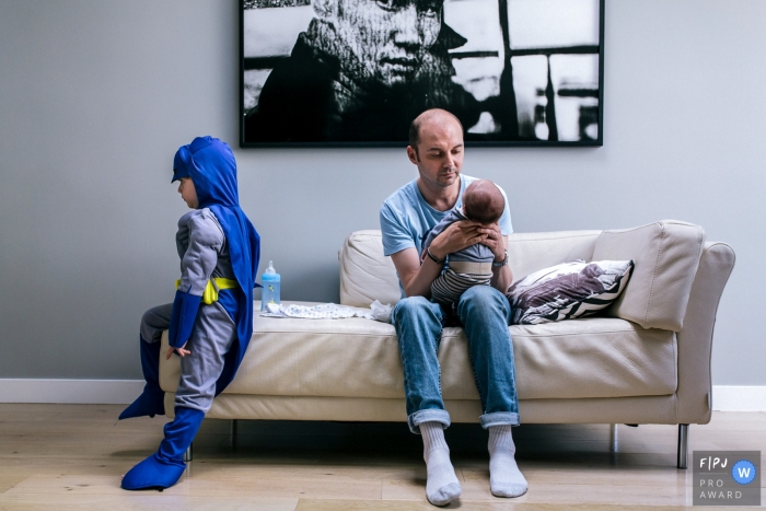 Photo by: <a href="https://www.fpja.com/documentary-family-photography-contests/marine-poron/paris/2081221">Marine Poron - France</a> A father holds his infant as his older son sits on the couch in a Batman costume in this photo by a Paris, France award-winning family photographer.