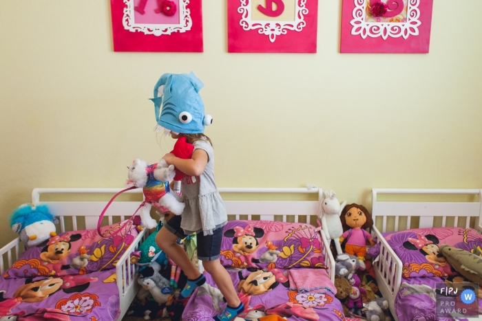 Photo by: <a href="https://www.fpja.com/documentary-family-photography-contests/danielle-daigle/phoenix/2082261">Danielle Daigle - United States</a> A little girl jumps on her bed with a toy fish on her head in this FPJA award-winning image captured by a Phoenix, AZ family photographer.