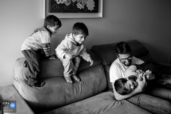 Un père joue avec ses trois fils sur un canapé sur cette photo enregistrée par un photographe de famille de style documentaire primé à Santa Catarina.