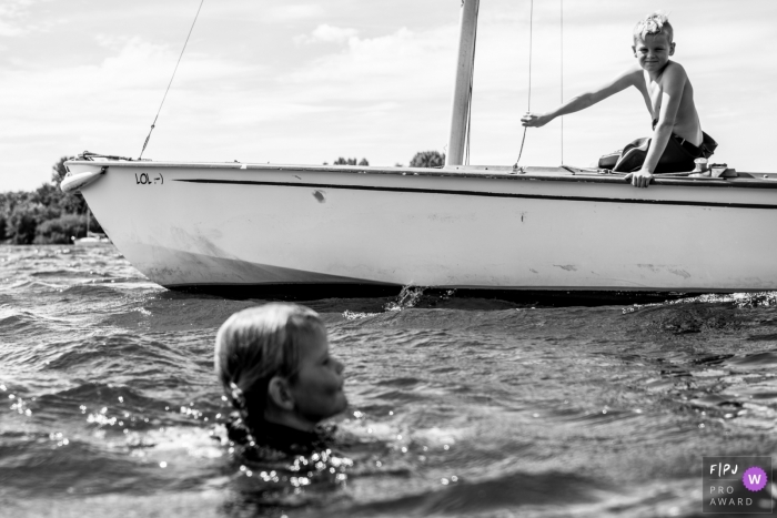 Un petit garçon regarde sa sœur nager dans l'eau depuis un voilier sur cette photo en noir et blanc réalisée par un photographe de la famille Eindhoven.