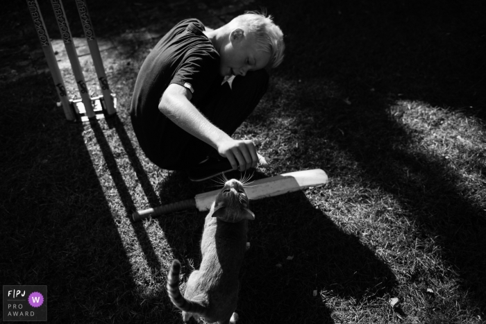 Un petit garçon prend une pause du cricket pour jouer avec un chat sur cette photo en noir et blanc réalisée par un photojournaliste de famille au style candide du Cambridgeshire, en Angleterre.