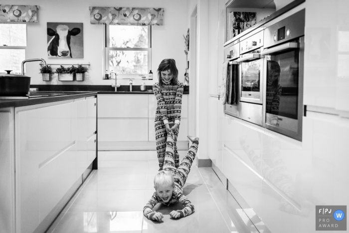 A girl pulls her brother along a tile floor in this black and white photo by a Cambridgeshire, English reportage family photographer. 