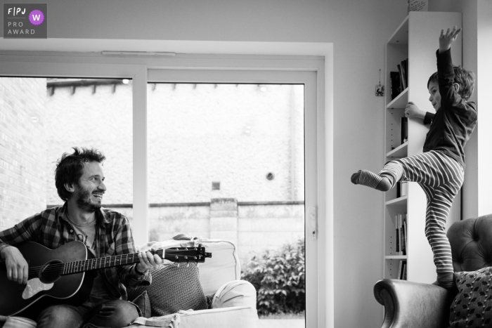 Un petit garçon danse sur une chaise pendant que son père joue de la guitare dans cette image familiale de style documentaire enregistrée par un photographe du Cambridgeshire, en Angleterre.