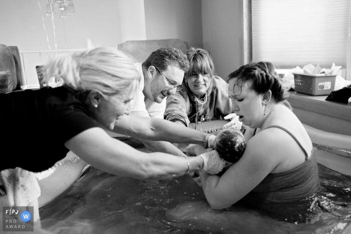 A mother, father, and nurses hold her newborn baby in a birthing tub after her at home water birth in this black and white photo by a Virginia family birth photographer. 