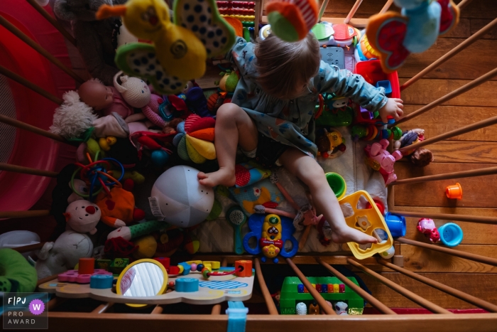Photo by: <a href="https://www.fpja.com/documentary-family-photography-contests/pieter-vandenhoudt/antwerpen/2084441">Pieter Vandenhoudt - Belgium</a> A little girl plays with the plethora of toys in her crib in this FPJA award-winning image captured by a Antwerpen family photographer.