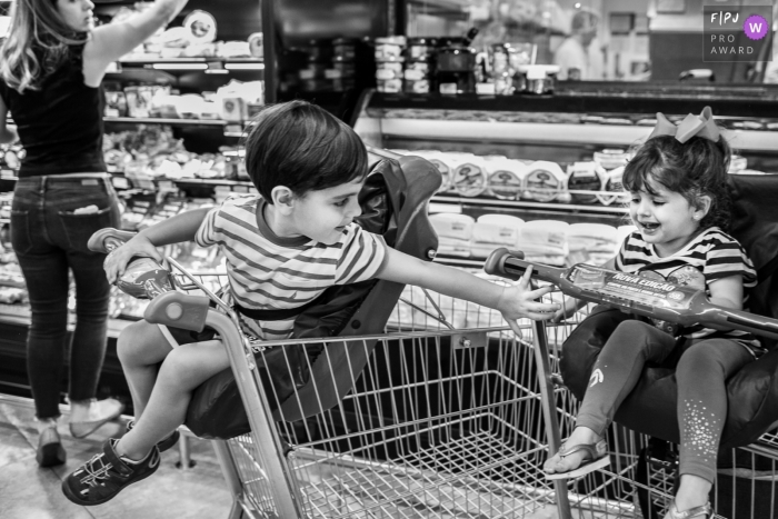 A little boy reaches back for his sister as they sit in grocery carts in this image created by a Minas Gerais family photographer.