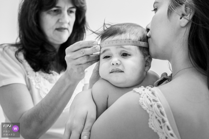 A mother kisses her baby as a woman measure's the infant's head in this award-winning photo by a Minas Gerais family photographer.