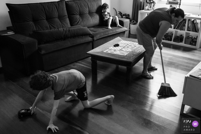 Une mère balaie le sol pendant que l’un de ses fils joue et l’autre assis sur un canapé dans cette image créée par un photographe de la famille Minas Gerais.