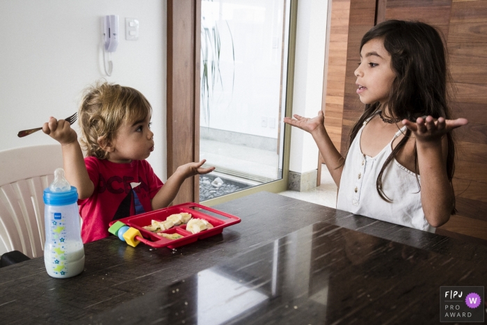 Photo by: <a href="https://www.fpja.com/documentary-family-photography-contests/leandro-malaquias/sete-lagoas/2084721">Leandro Malaquias - Brazil</a> A little girl and boy shrug at each other while the boy eats lunch in this picture captured by a Minas Gerais family photojournalist.