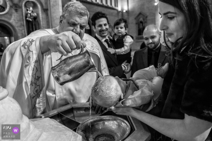 Photo by: <a href="https://www.fpja.com/documentary-family-photography-contests/adeline-leonti/montreal/2085056">Adeline Leonti - Canada</a> A mother holds her baby as a priest performs the baptism in this Family Photojournalist Association contest awarded photo created by a Montreal, Quebec family photographer.