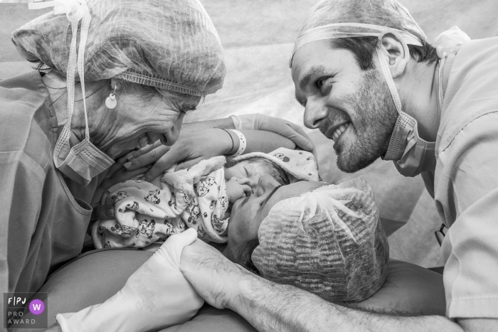 A woman's mother and husband hug her as she holds her newborn in the hospital in this black and white picture captured by a Rio de Janeiro, Brazil birth and newborn photographer. 