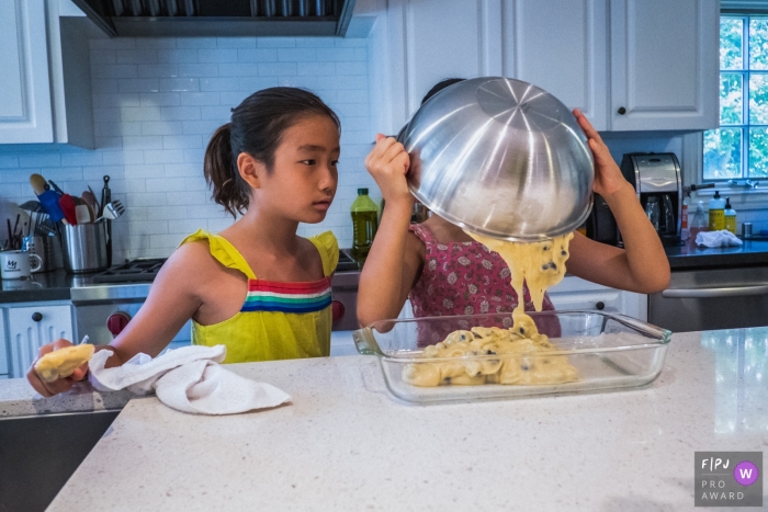 A girl pours batter into a baking pan as her sister watches in this Family Photojournalist Association awarded photo by a Los Angeles, CA documentary family photographer.