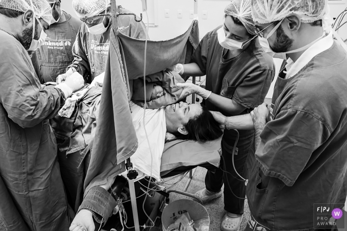 Photo by: <a href="https://www.fpja.com/documentary-family-photography-contests/gisele-soares/rio-de-janeiro/2085811">Gisele Soares - Brazil</a> Nurses hold a baby up for its mother to see immediately following her hospital birth in this black and white documentary picture by a Rio de Janeiro, Brazil birth photographer.