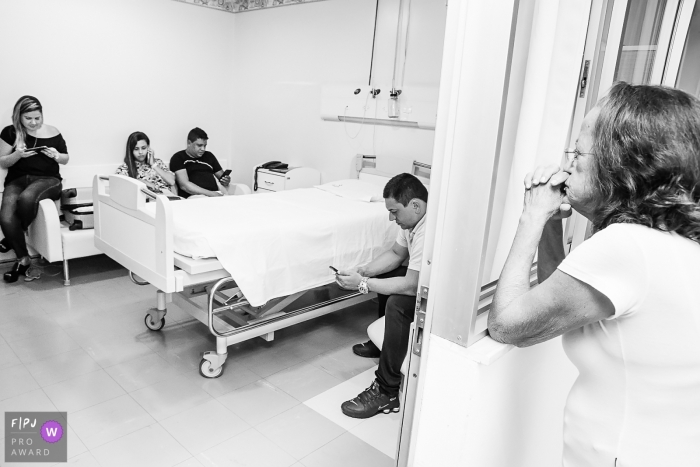 A family waits in a hospital room as one woman prays in this black and white image recorded by a Rio de Janeiro, Brazil birth photographer. 