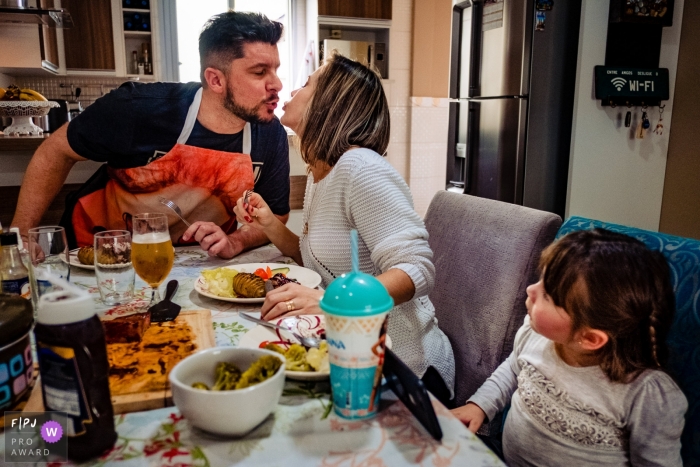 Photo by: <a href="https://www.fpja.com/documentary-family-photography-contests/rafael-rosa/rio-grande-do-sul/2085946">Rafael Rosa - Brazil</a> A husband and wife kiss at the dinner table as their daughter looks at them in this image created by a Rio Grande do Sul, Brazil family photographer.