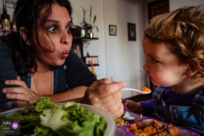 A mother makes faces as she tries to get her young daughter to eat in this photo recorded by a Rio Grande do Sul, Brazil award-winning, documentary-style family photographer. 