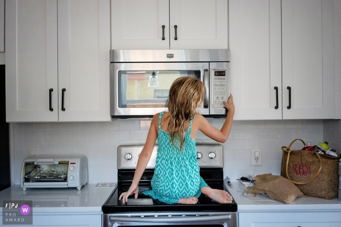 Photo by: <a href="https://www.fpja.com/documentary-family-photography-contests/julie-ambos/key-west/2086066">Julie Ambos - United States</a> A young girl sits on the stove to reach the microwave in this Family Photojournalist Association contest awarded photo created by a Key West, FL family photographer.