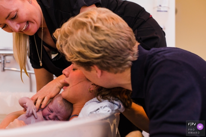 Two women support a woman in a birthing tub as she holds her newborn for the first time in this photo composed by a Copenhagen documentary birth photographer.