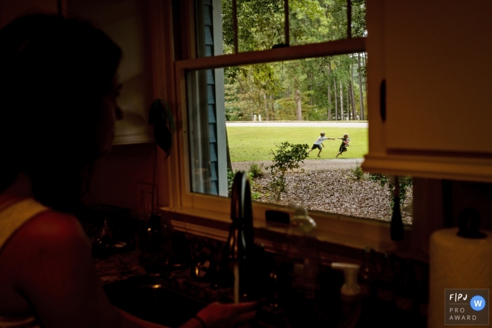 A mother watches her children play through the kitchen window in this documentary-style family photo captured by a Key West, FL photographer. 