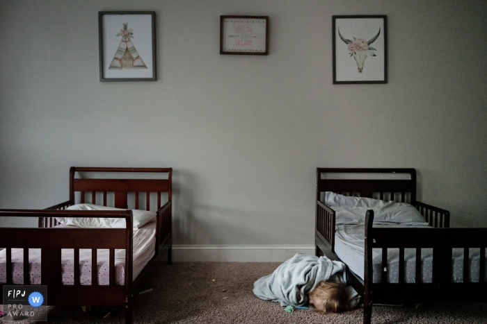 A young child sleeps wrapped in a blanket on the ground right next to a bed in this award-winning photo by a Key West, FL family photographer.