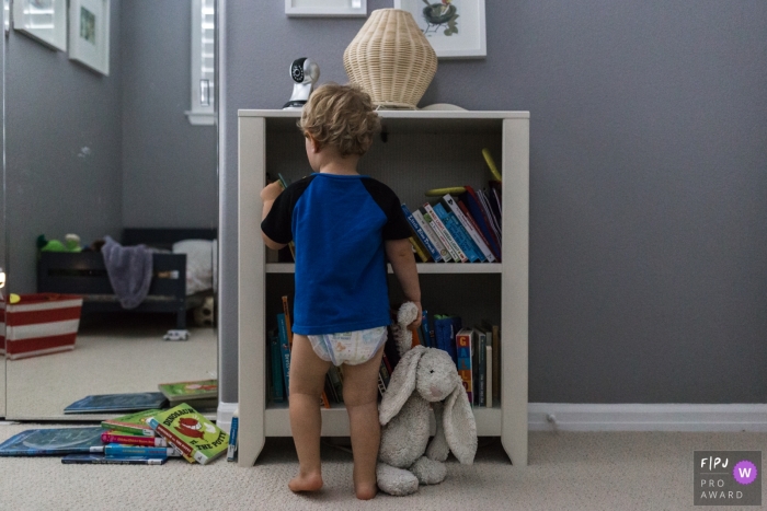 Photo by: <a href="https://www.fpja.com/documentary-family-photography-contests/melissa-mcclure/los-angeles/2086331">Melissa McClure - United States</a> A little boy looks through a bookshelf while holding his stuffed rabbit in this picture captured by a Los Angeles, CA family photojournalist.