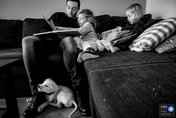 North Rhine-Westphalia documentary family photographer captured this black and white photo of a family enjoying storytime on the couch while the puppy lays on the floor below