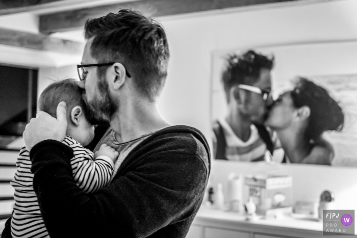Photo by: <a href="https://www.fpja.com/documentary-family-photography-contests/katrin-k%C3%BCllenberg/dusseldorf/2086516">Katrin Küllenberg - Germany</a> This black and white photo of a father kissing his baby in front of a portrait of him kissing his partner was captured by a North Rhine-Westphalia documentary family photographer