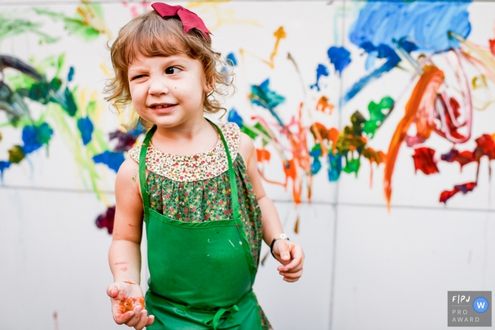 Photo by: <a href="https://www.fpja.com/documentary-family-photography-contests/gustavo-medeiros/rio-de-janeiro/2086546">Gustavo Medeiros - Brazil</a> Rio de Janiro documentary family photographer captured a this photo of a paint covered little girl winking while wearing a green apron and a red bow in front of a paint smeared wall