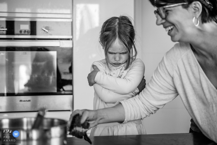 Une photographe de famille de documentaire Auvergne-Rhône-Alpes a capturé cette photo en noir et blanc d'une jeune fille qui fait la moue dans la cuisine pendant que sa mère nettoie