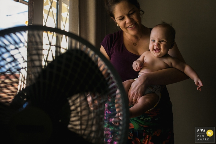 A mother holds her baby in front of a fan in this documentary-style family image recorded by a Minas Gerais photographer. 