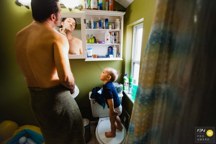 Photo by: <a href="https://www.fpja.com/documentary-family-photography-contests/pamela-anticole/pittsburgh/2089746">Pamela Anticole - United States</a> A baby boy stands on a toilet seat as he watches his father shave in this FPJA award-winning picture by a Pittsburgh family photographer.