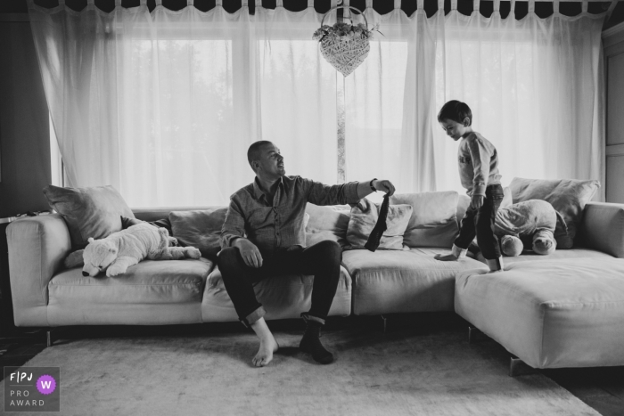 Photo by: <a href="https://www.fpja.com/documentary-family-photography-contests/alice-franchi/pistoia/2089761">Alice Franchi - Italy</a> A boy stands on a couch as his father takes off his socks in this documentary-style family photo captured by a Pistoia photographer.